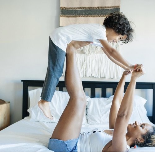 A woman lying on a mattress in a bedroom, on her back while holding a little boy up on her feet and hands in the air