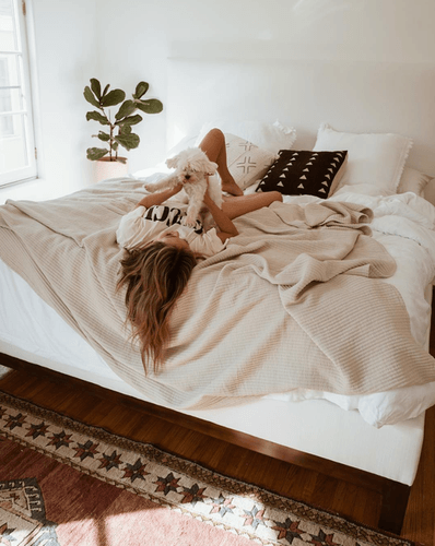 A bedroom setting with a potted plant, rug, mattress, foundation; a woman lying on the bed holding her dog
