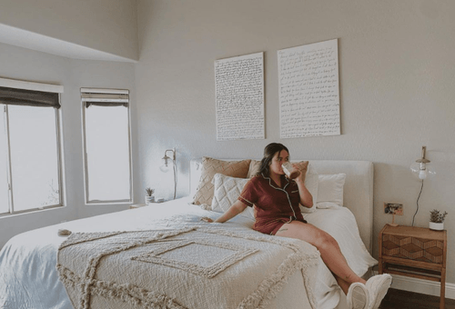 Woman dressed in pajamas and slippers sitting on dressed bed sipping out of mug