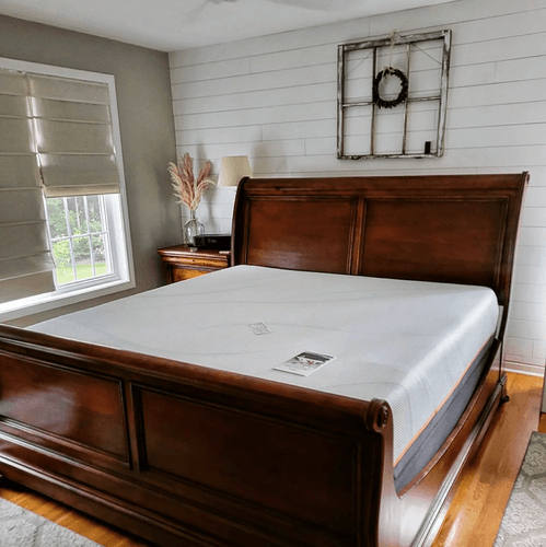 Mattress on a wooden bed frame in a decorated room with white walls, bedside table, and tan curtains