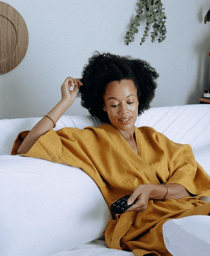 Woman dressed in yellow robe leaning on pillows on mattress holding remote to Tempur-Pedic Ergo Smart Base