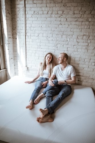 A smiling couple sitting on a Tempur-Cloud mattress in a well lit room